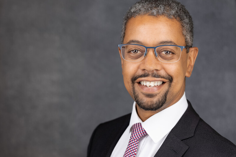 Professional headshot of a man wearing glasses, dark blazer, and striped tie against neutral gray background.