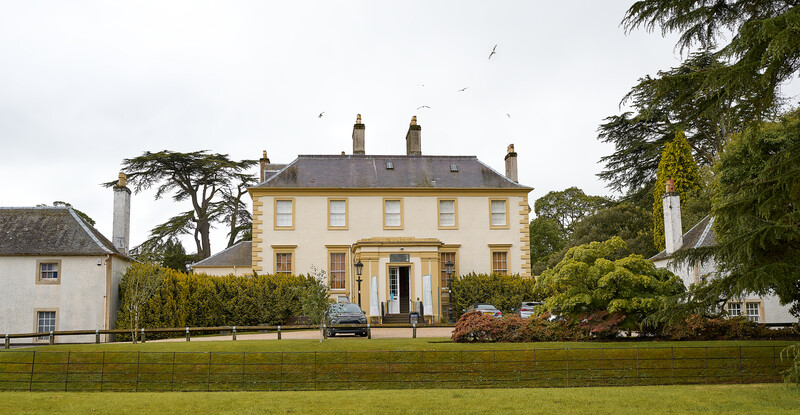 Pale yellow Georgian mansion with slate roof framed by mature trees and manicured lawn.