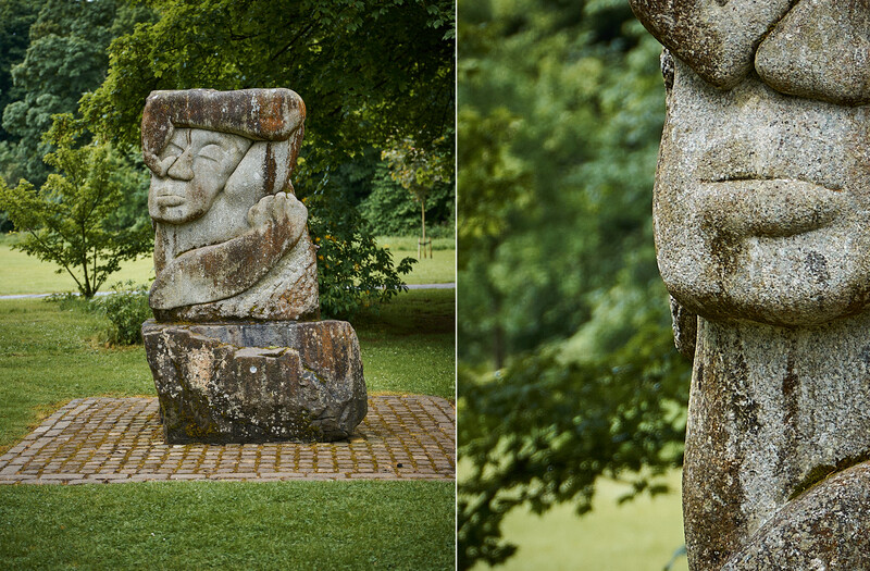 Weathered stone sculpture of a stylized human head on a brick plaza, surrounded by green lawn and trees.