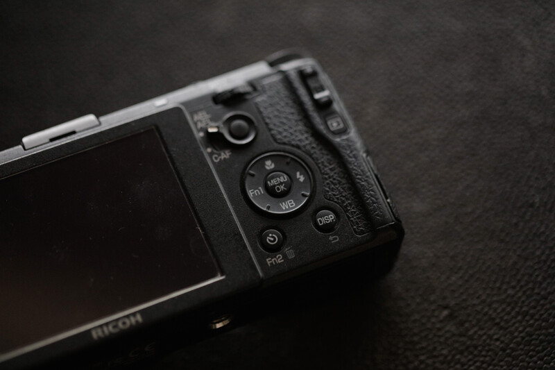 Ricoh compact camera with control dial and buttons visible on dark textured surface.