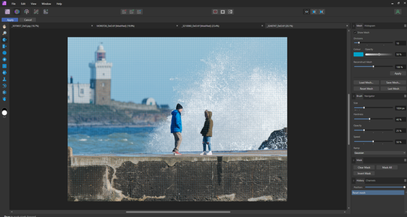 Two people standing on a stone wall watching large waves crash against the barrier, with a coastal city and lighthouse visible across the water.