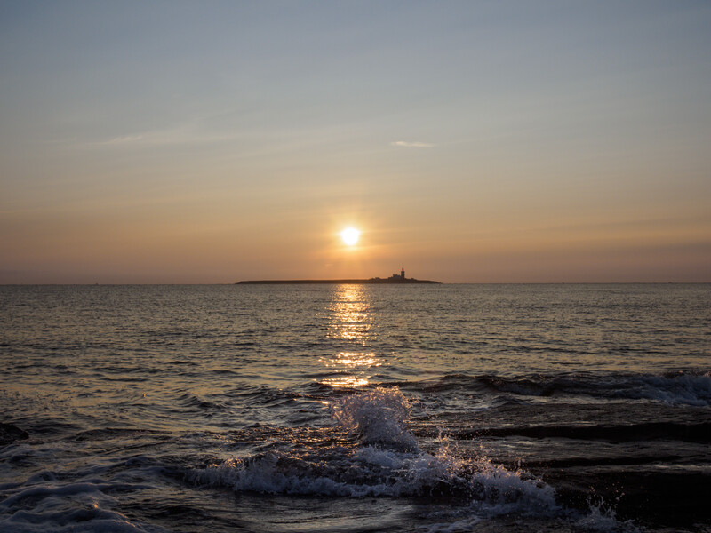 Sun setting over calm ocean with waves breaking on shore and silhouetted landmass on horizon.