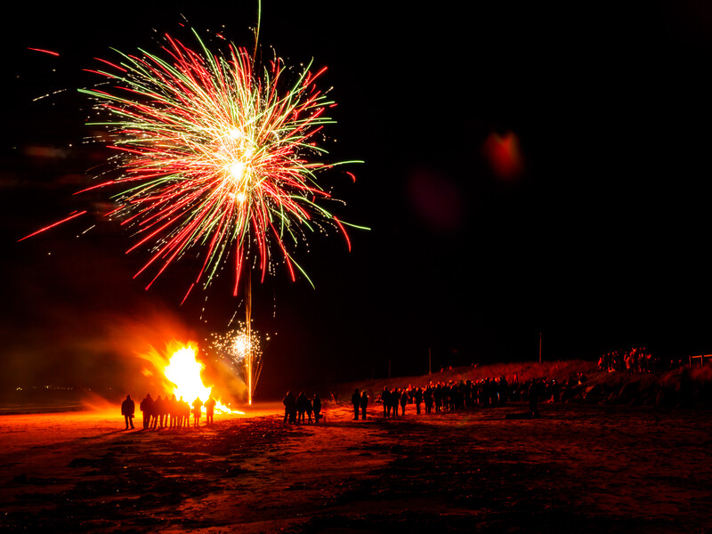 Fireworks bursting overhead above a nighttime beach bonfire with silhouetted spectators gathered around.