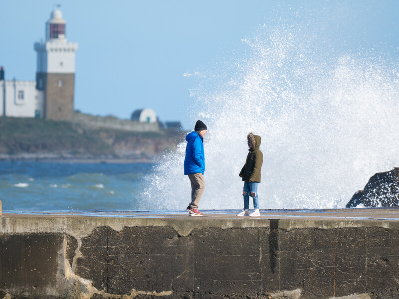 Two children standing on a stone seawall as a large wave crashes behind them, with a lighthouse visible on a distant headland.