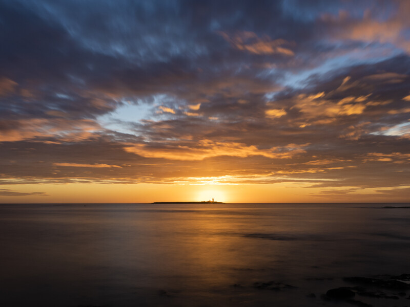 Golden sunset over calm ocean water with dramatic cloud formations and distant landmass silhouette.
