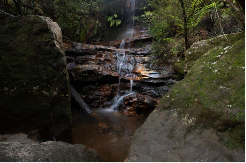 Multi-tiered waterfall cascading over dark rocks surrounded by moss-covered stone walls and lush green vegetation.