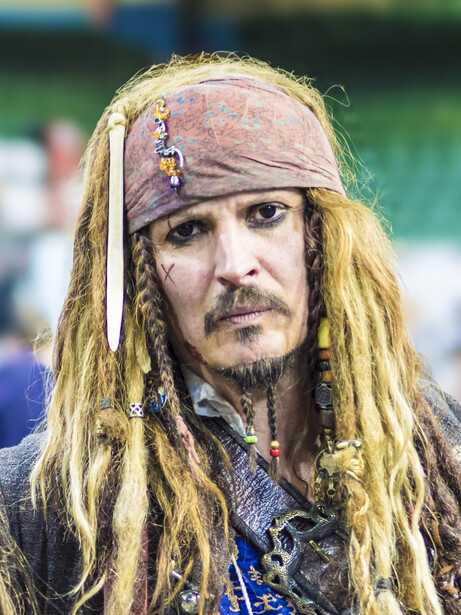Person with long blonde dreadlocks and face paint wearing a headscarf and ornate costume.