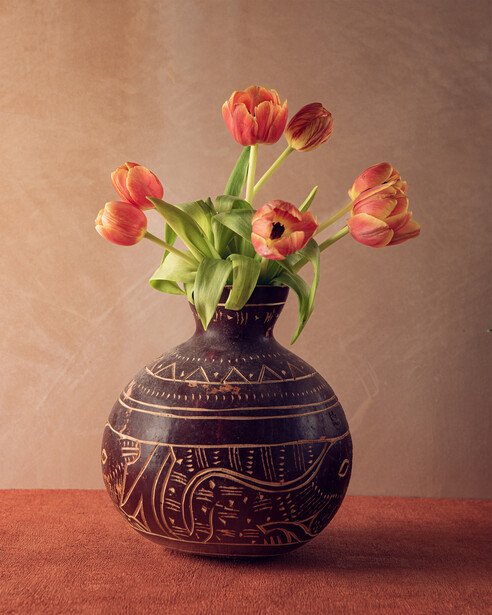 Red and orange tulips arranged in a dark decorative ceramic vase against a warm neutral background.