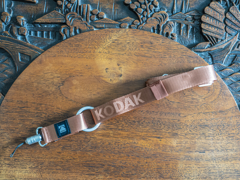 Kodak camera strap with metal fittings lying diagonally on a wooden surface with carved decorative background.