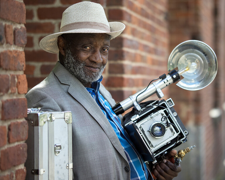 Photographer holding a large format camera and studio flash against a brick wall background.