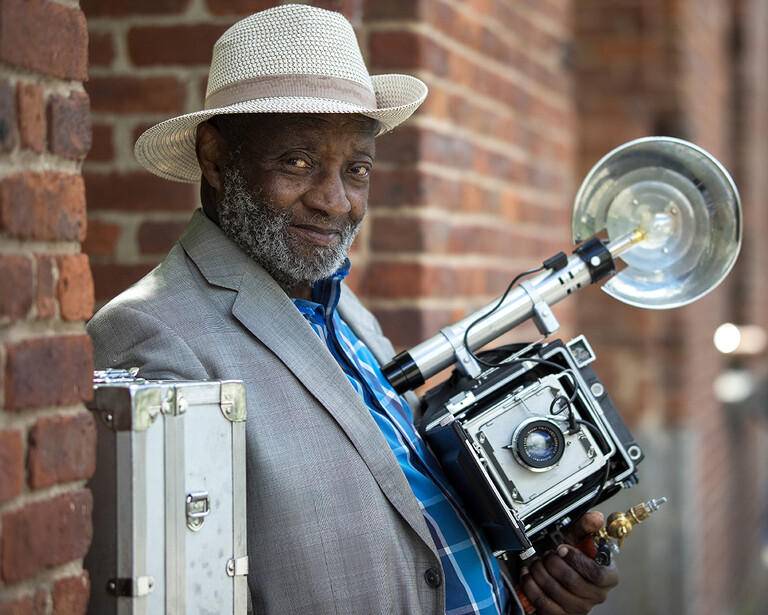 Photographer holding a large format camera with flash attachment against a brick wall backdrop.