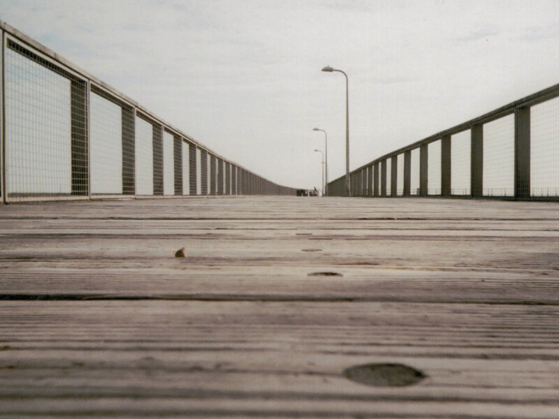 Low-angle perspective down an empty wooden boardwalk or pier between metal railings stretching toward the horizon.