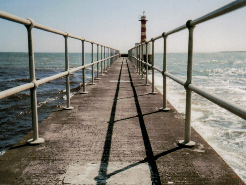 Long concrete pier with metal railings extending into calm ocean waters, lighthouse visible in distant background.