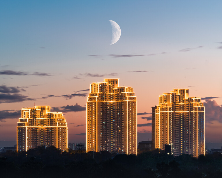 Illuminated residential high-rises at dusk with a crescent moon in a twilight sky.