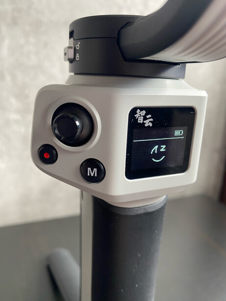 Close-up of a camera gimbal head with white body, black pan handle, and control panel with joystick and buttons.