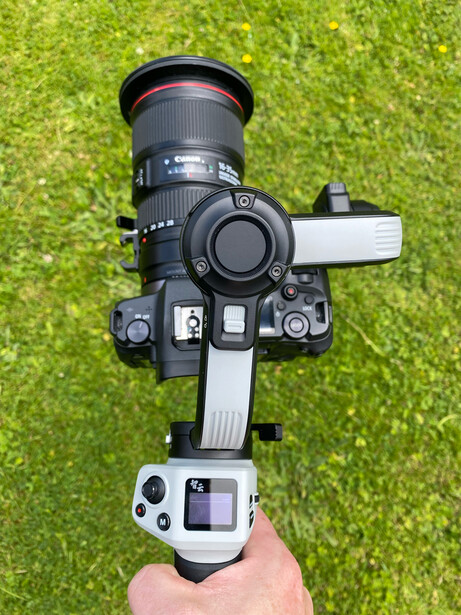 Overhead view of a mirrorless camera with telephoto lens mounted on a white gimbal head, held in a hand against green grass background.