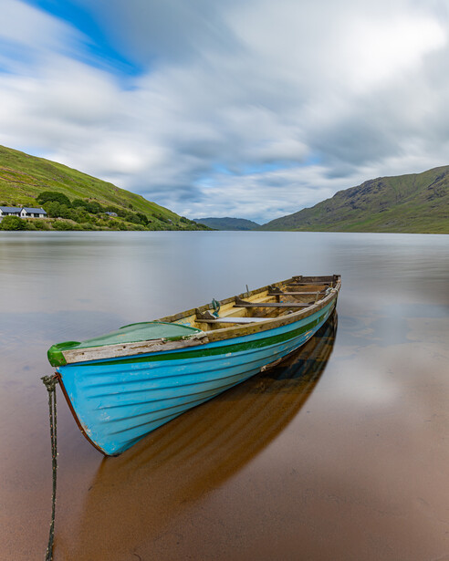Blue and green wooden rowboat anchored in calm water with green hillsides and mountains in the background.
