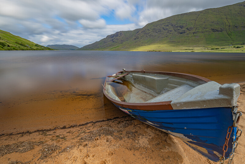 Blue and white wooden rowboat beached on sand beside a calm lake with green mountains beyond.