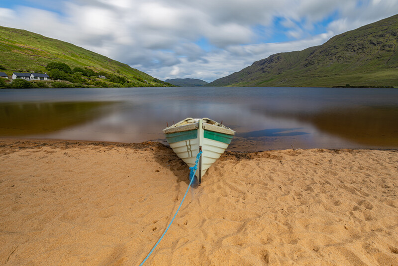 Green and blue rowboat beached on sand at the shore of a calm mountain lake.