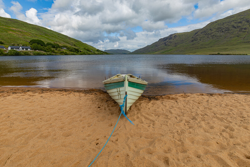 Green and blue wooden boat beached on sand at the edge of a calm lake, with mountains and a small cottage visible across the water.