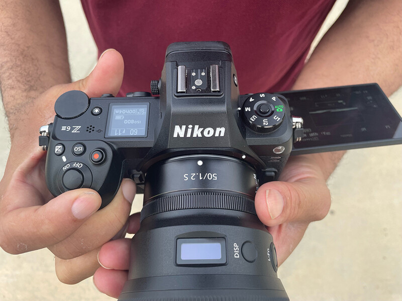 Hands holding a Nikon mirrorless camera with 50mm lens mounted, showing top controls and articulating screen.