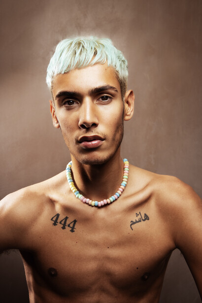 Shirtless man with platinum blonde hair and chest tattoos against neutral brown background.