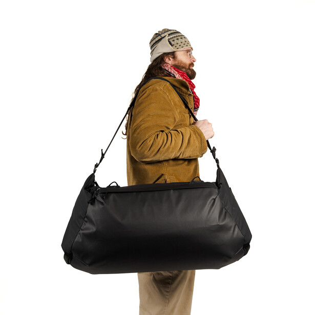 Model wearing brown jacket and beanie carrying oversized black duffle bag against white background.
