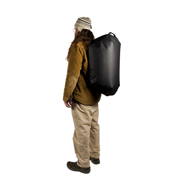 Person wearing a mustard jacket and beige pants carrying a large black cylindrical backpack.
