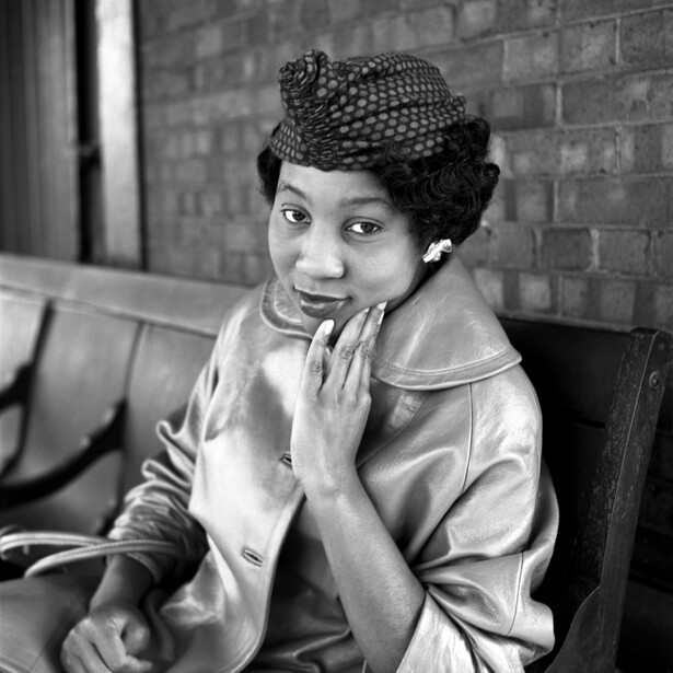Woman in vintage clothing and netted hat, posed against brick wall in black and white portrait.