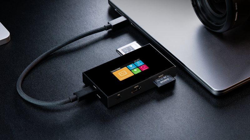 Portable external SSD with colorful menu interface connected to laptop and camera via cables on dark desk.