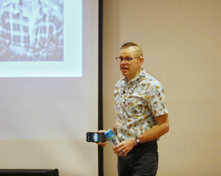 Man in patterned shirt and glasses presenting at a seminar, holding a device while standing before a projection screen.
