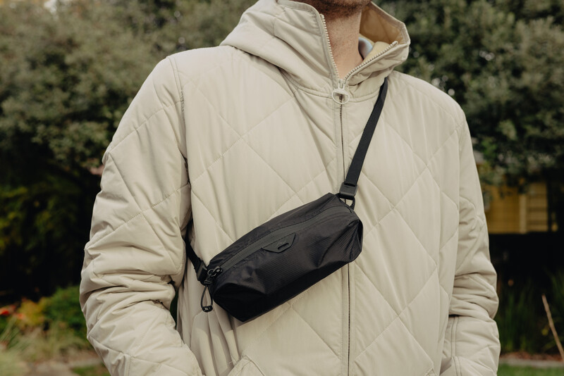 Man wearing beige quilted puffer jacket with black crossbody sling bag against blurred green foliage background.