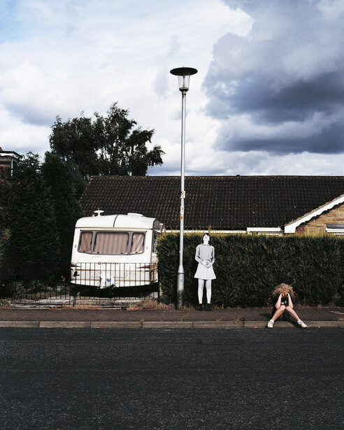 Vintage caravan parked beside a residential fence with two figures in the foreground on an overcast day.
