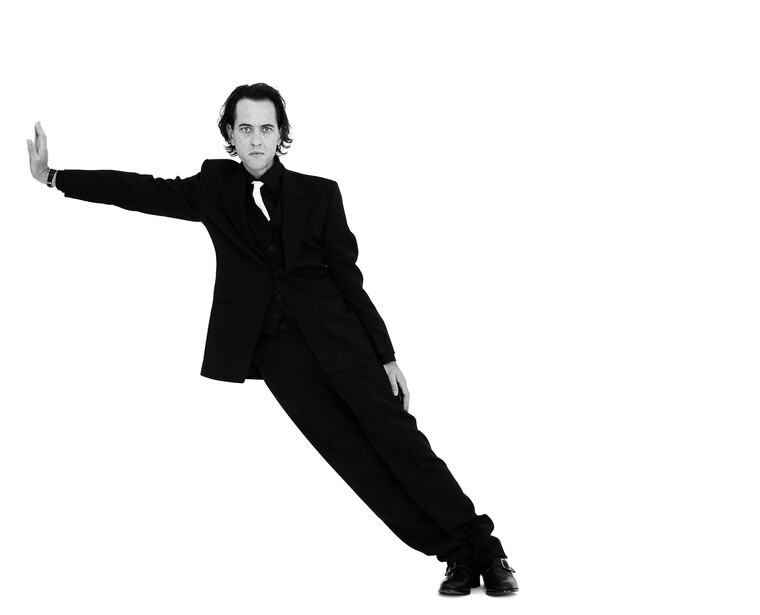 Man in black suit leaning dramatically to the side against white background.