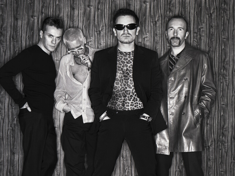 Four men in dark clothing posed against a textured backdrop in black and white.