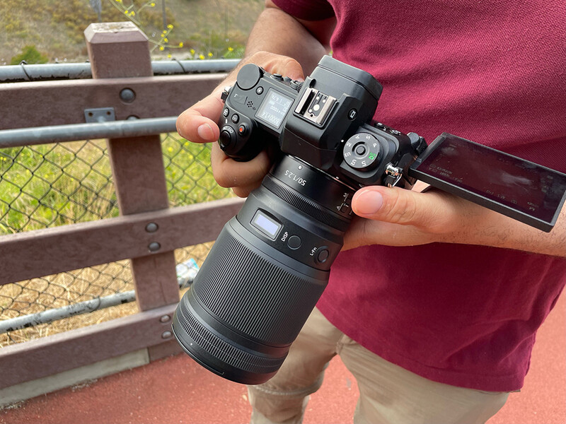 Photographer holding a mirrorless camera with telephoto lens attached, shot outdoors against a wooden fence.