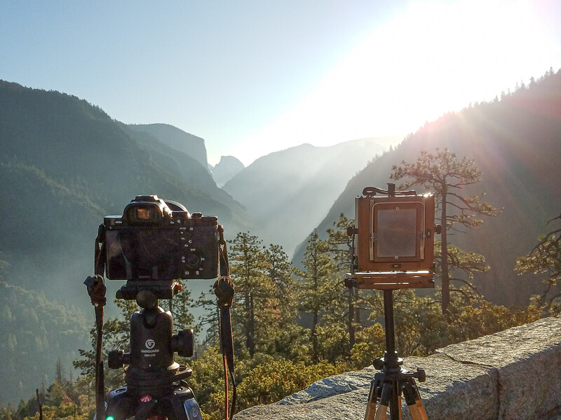 Two tripod-mounted cameras positioned on a stone overlook facing a sunlit mountain valley surrounded by forested peaks.