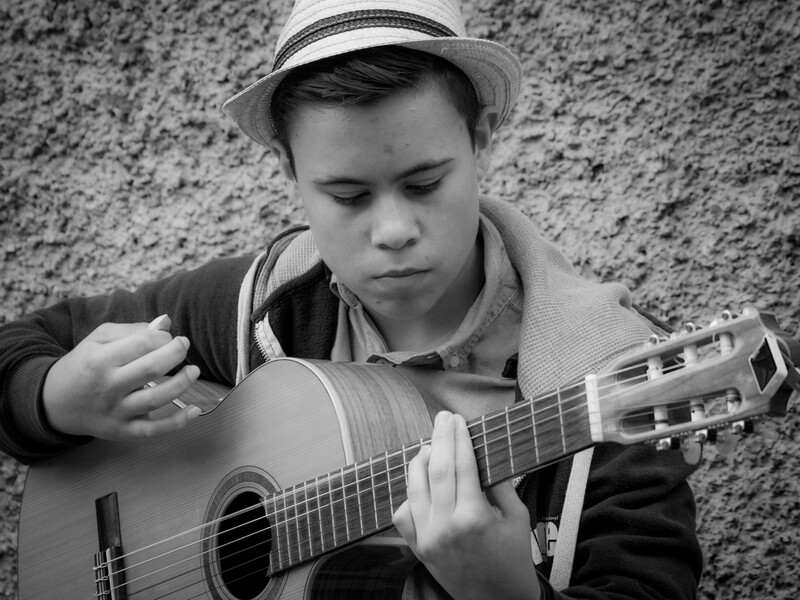 Young boy playing classical guitar against a textured concrete wall, wearing a hat and focused on his instrument.