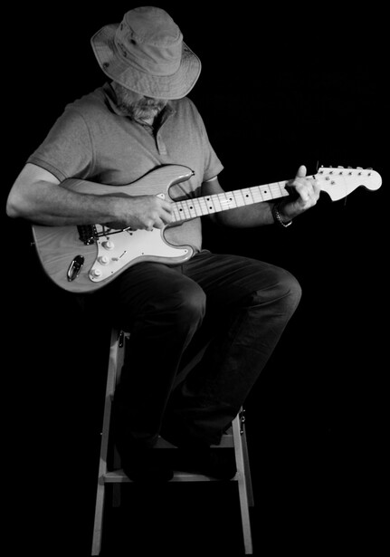Musician seated on a stool playing an electric guitar against a black background.