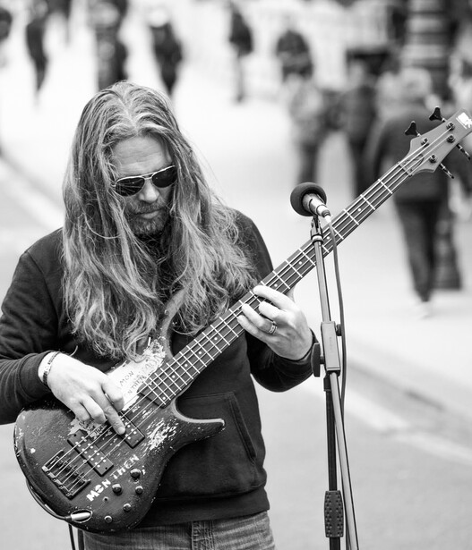 Long-haired musician wearing sunglasses plays bass guitar at a microphone during a street performance.