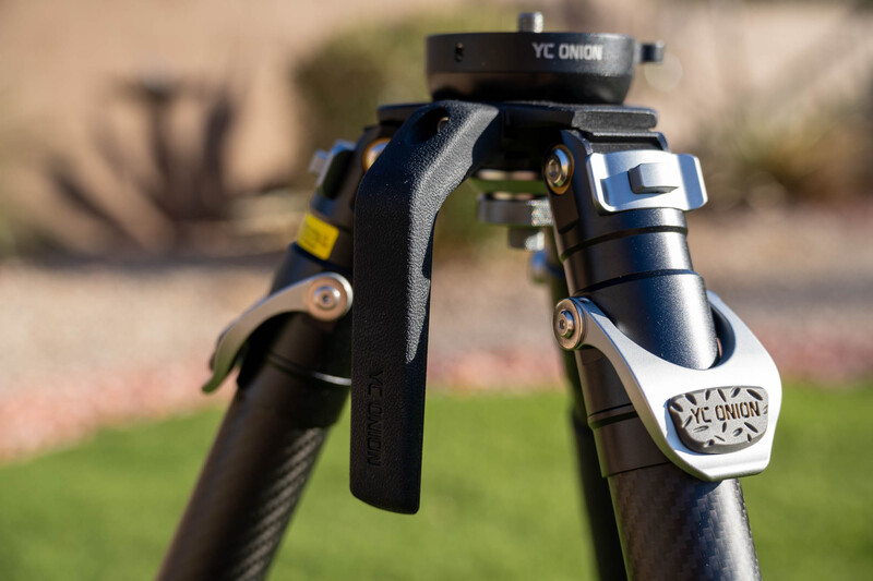 Close-up of a camera tripod head with gray straps and metal components against a blurred outdoor background.