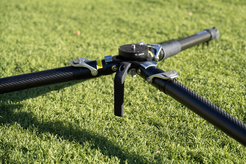 Tripod head with pan-tilt mechanism mounted on grass, shown from above with extended arms.