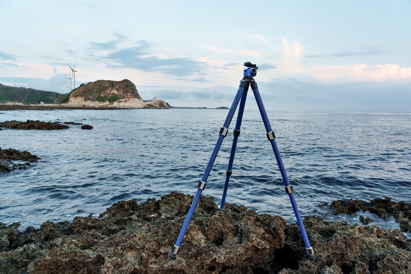 Blue tripod positioned on rocky coastal outcrop with island and calm sea in background.