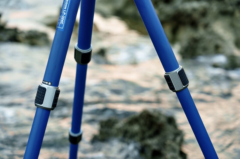 Blue tripod legs with metal connectors in sharp focus, blurred rocky landscape and water behind.