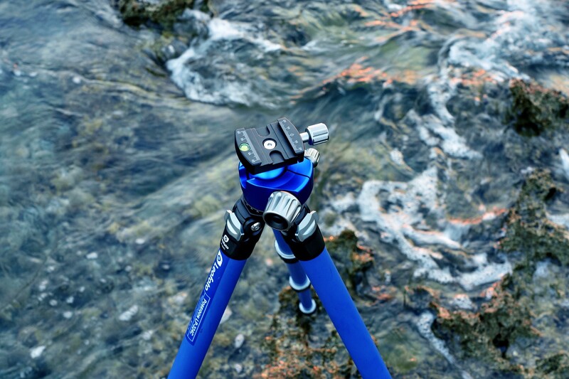 Blue spearfishing gun with black trigger mechanism pointed at rocky seafloor with shallow water and algae-covered rocks.