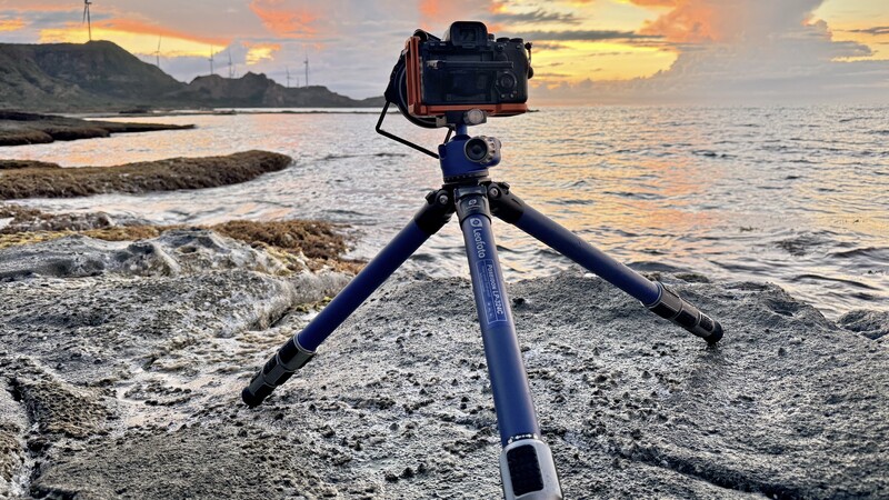 DSLR camera mounted on blue tripod positioned on rocky coastline at sunset.