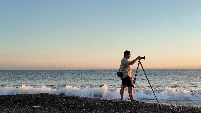 Photographer on a pebbly beach at sunset, using a tripod-mounted telephoto lens to capture the ocean and waves.