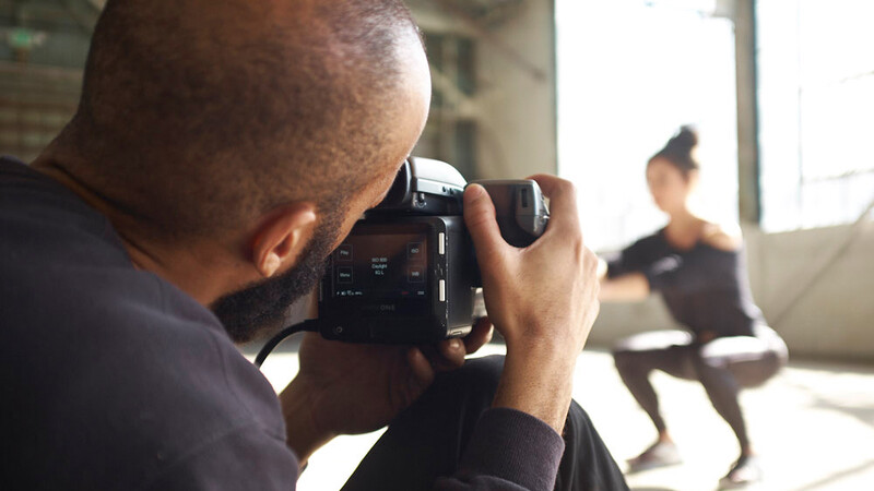 Photographer holding a professional video camera, filming a person dancing in a studio with natural window light.