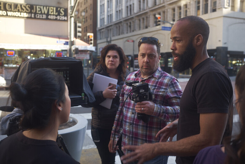 Filmmaking crew members gathered on an urban street discussing production, with one holding a professional camera.