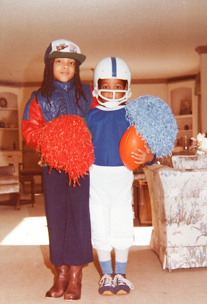 Two people in a living room wearing sports uniforms and foam accessories, posed for a casual portrait.